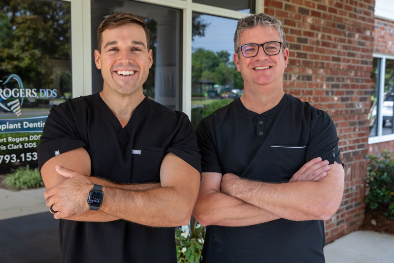 Dr. Chris Clark and Dr. Robert Rogers, trusted family dentists at Rogers Clark & Associates in Concord, NC, standing outside their dental office smiling with arms crossed.